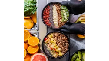 Two smoothie bowls topped with sliced fruits, granola, chia seeds, and nuts on a kitchen counter with fresh oranges.