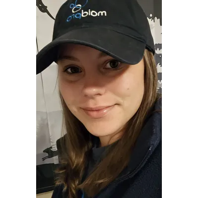 Close-up of a smiling woman wearing a black biqbiom cap and dark jacket with long brown hair.
