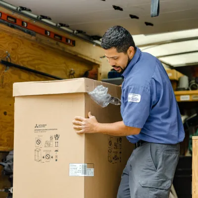Delivery worker unloading large sealed cardboard box from a truck amid tools and equipment.