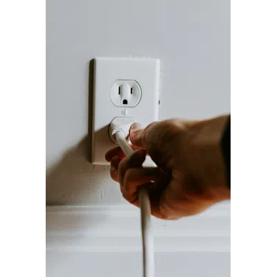 Hand plugging a white electrical cord into a wall outlet on a white painted wall with baseboard.