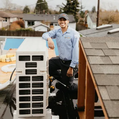 Smiling HVAC technician in uniform kneeling on a roof next to an air conditioning unit with tools in daylight.