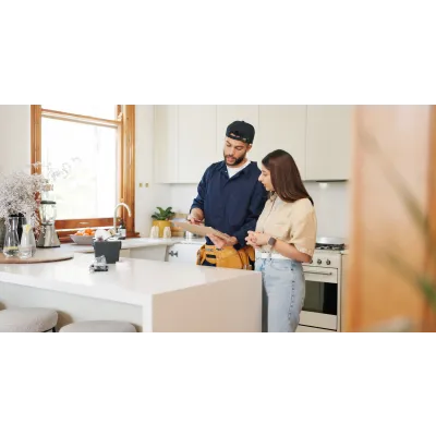 Man in work uniform explaining repairs to woman in modern kitchen with white countertops and natural light