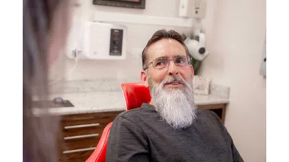 Bearded man with glasses sitting on a red dental chair talking in a dental office setting.
