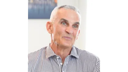 Smiling elderly man with gray hair wearing a black and white checkered shirt in a bright indoor setting.