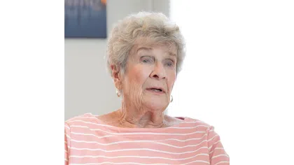 Elderly woman with gray hair wearing a pink striped shirt, speaking in a softly lit room with neutral background.
