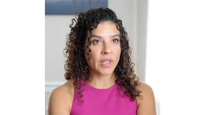 Woman with curly hair wearing a bright pink sleeveless top speaking indoors with a neutral background.
