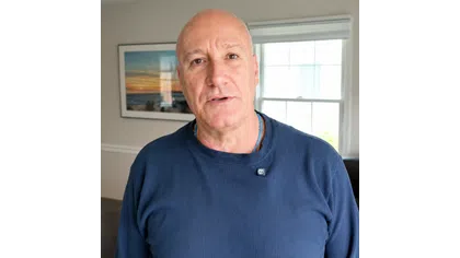 Middle-aged bald man wearing a navy blue long-sleeve shirt standing indoors with a window and wall art in the background