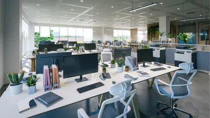Modern open-plan office with desks, computers, ergonomic chairs, plants, and ample natural light from large windows.