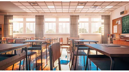 Sunlit empty classroom with wooden desks, chairs, large windows, and a chalkboard on the wall.