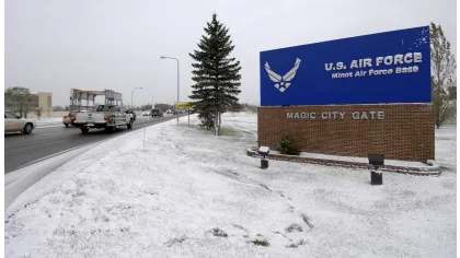Snow-covered entrance sign for U.S. Air Force Minot Air Force Base Magic City Gate with vehicles driving on road.