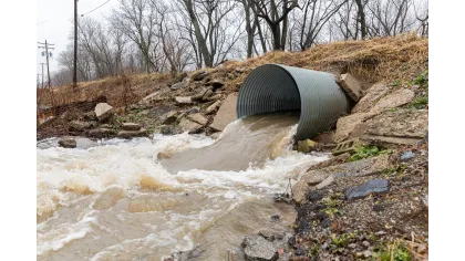 Floodwater rushing out of a large drainage pipe into a creek with bare trees and rock embankment in early spring.