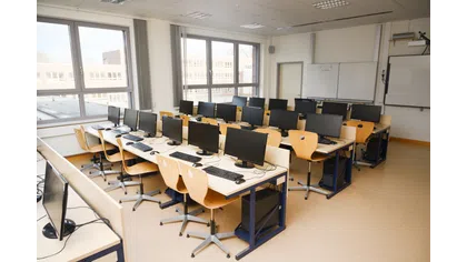 Empty computer lab with multiple desktop computers, wooden chairs, large windows, and whiteboards in a classroom.