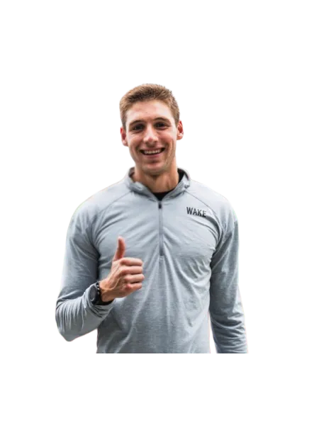 Smiling young man in gray WAKE sweatshirt giving thumbs up, standing confidently with black watch and striped pants.