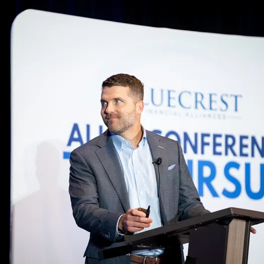 Man in gray suit speaking at Bluecrest Financial Alliance conference podium with presentation screen behind him