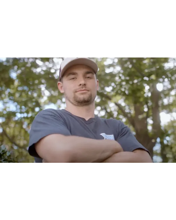 Young man wearing a cap and navy blue shirt stands with arms crossed outdoors with blurred green trees in background.
