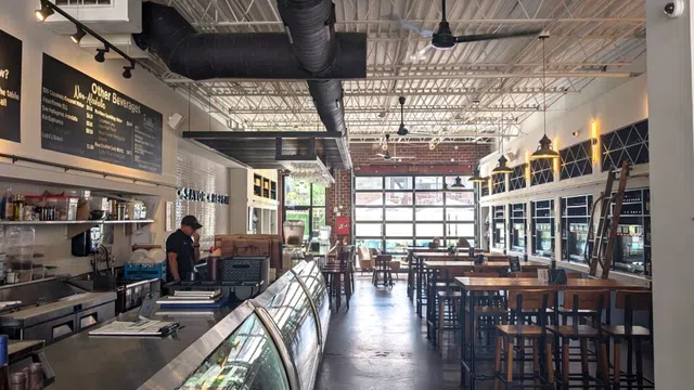 Modern cafe interior with high ceilings, wooden tables and chairs, glass display case, and menu boards.