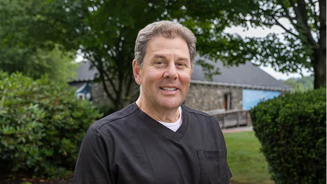 Middle-aged man in black scrub top smiling outdoors with trees and a rustic building in the background