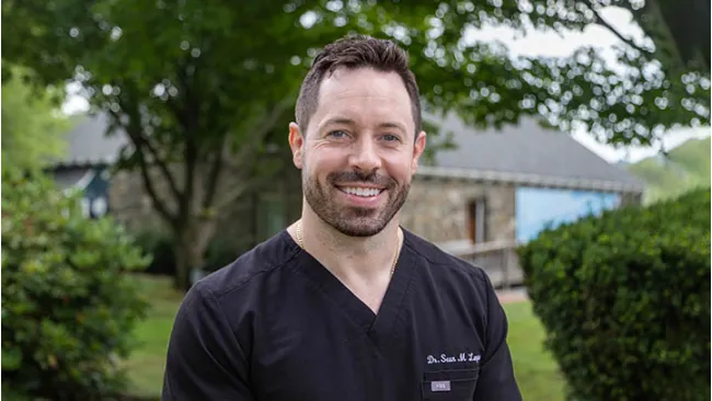 Smiling man in black medical scrubs standing outdoors with trees and a stone building in the background