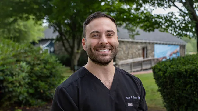 Smiling man in a black scrub top stands outdoors by greenery and a stone building on a bright day.