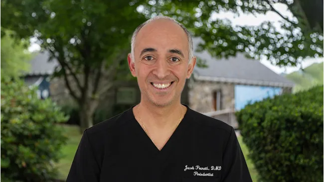 Smiling middle-aged male doctor in black scrubs standing outdoors with trees and a building in the background.