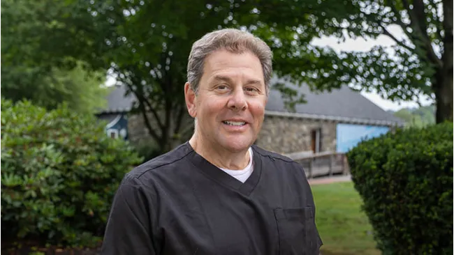Middle-aged man in black scrub top smiling outdoors with trees and a stone building in the background
