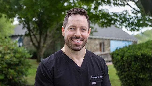 Smiling man in black medical scrubs outdoors with greenery and a stone building in the background