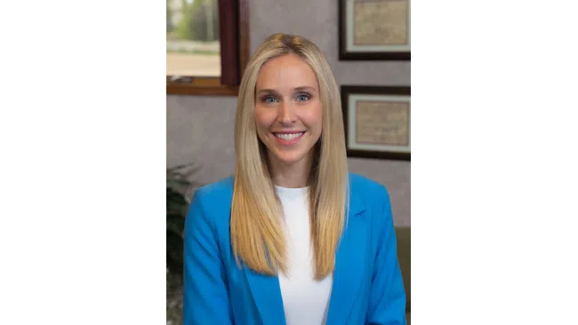 Smiling blonde woman in a blue blazer and white shirt posing indoors with framed pictures in the background.