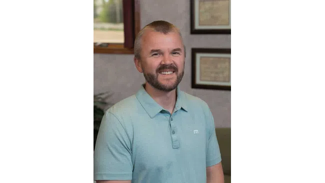 Smiling man with beard wearing a light blue polo shirt indoors with framed pictures on the wall behind him.