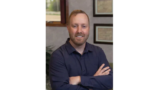 Smiling man with beard and short hair in navy shirt standing with arms crossed indoors by framed certificates.