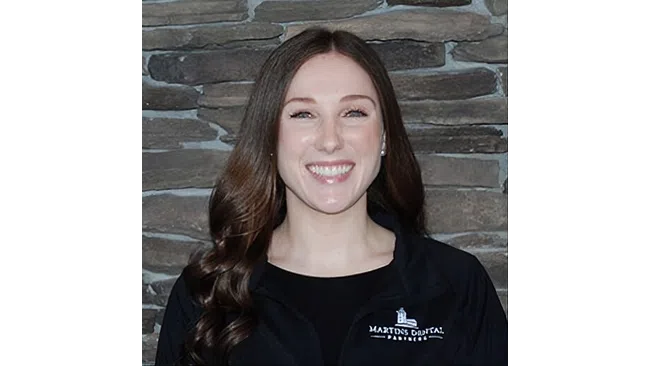 Smiling woman with long brown hair wearing a black jacket standing in front of a stone wall