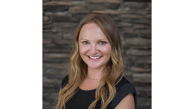 Portrait of a smiling woman with long blonde hair wearing a black top against a stone wall background