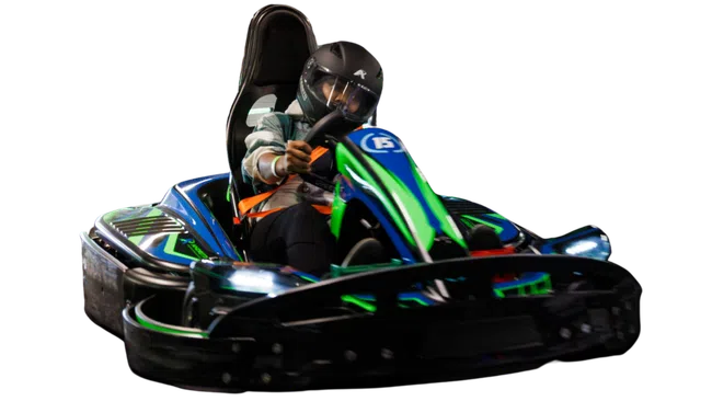 Person wearing a helmet driving a blue and green go-kart racing vehicle at speed indoors.