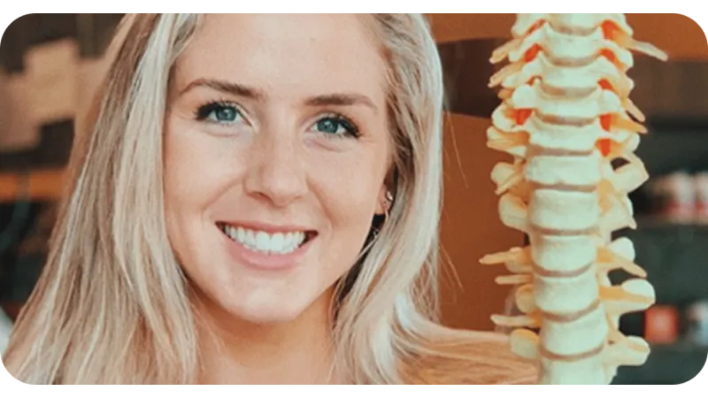Smiling woman with blonde hair holding a model of a human spine against a blurred background