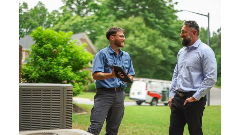 HVAC technician consulting with homeowner outside near air conditioning unit and service van in a residential neighborhood.