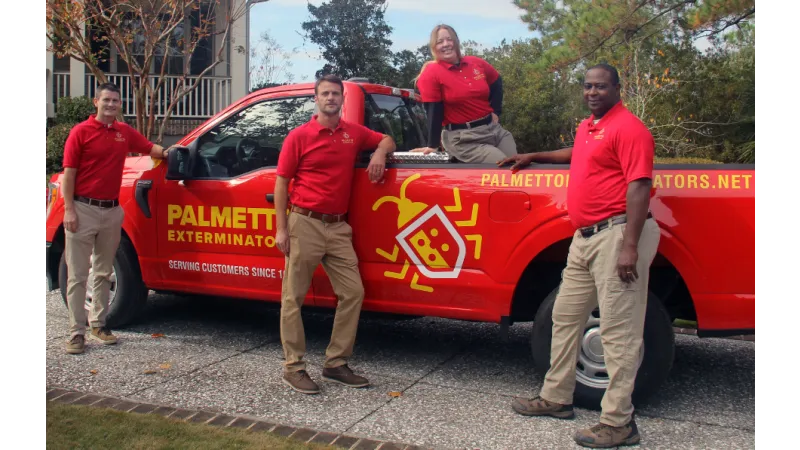 Four pest control workers in red shirts posing with a red Palmetto Exterminators truck outdoors.