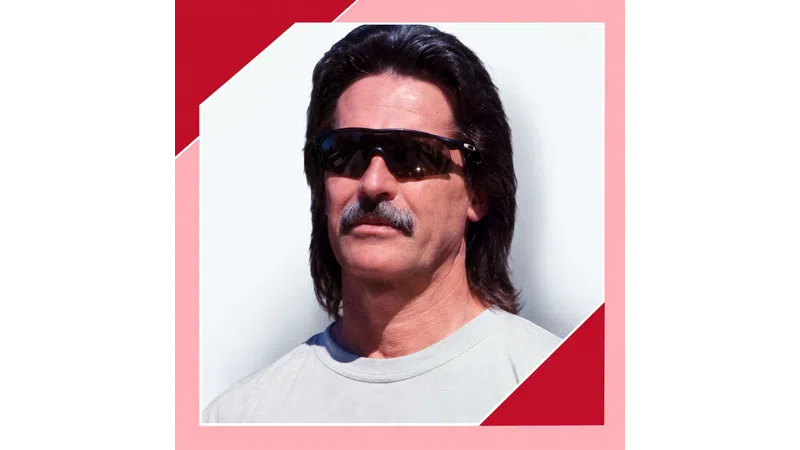 Man with mullet hairstyle, mustache, and dark sunglasses wearing a light gray shirt against white and red background.