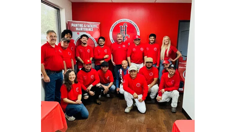 Group of employees in red shirts posing inside a Harrison Contracting Company office with branding banners.