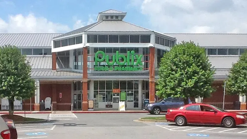 Publix supermarket front entrance with green signage, cars parked, and trees on a sunny day.