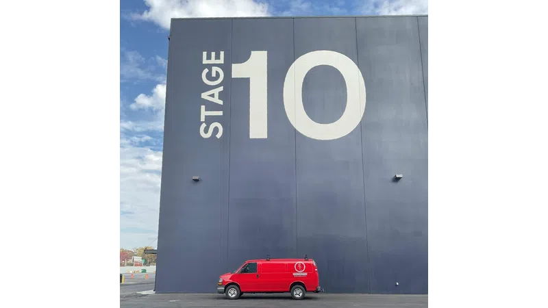 Large dark building labeled Stage 10 with a red van parked in front under a cloudy blue sky