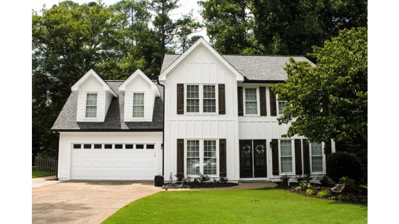 Two-story white house with black shutters, attached garage, and well-maintained green lawn in a wooded neighborhood.