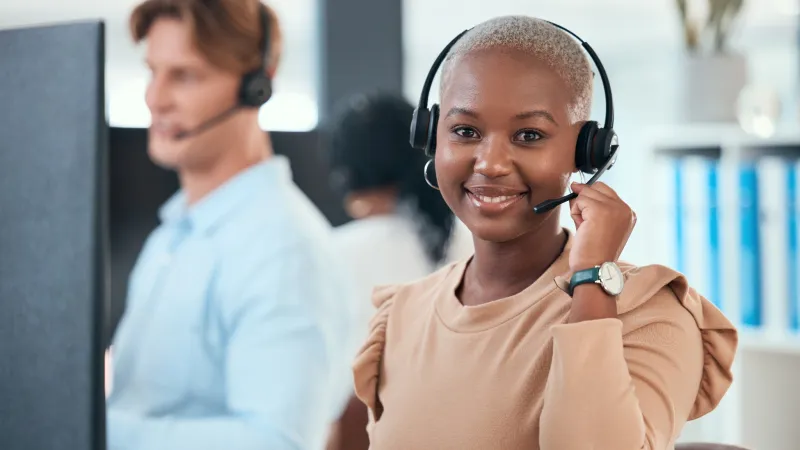 Smiling customer service representative wearing headset in a busy office with colleagues working in background