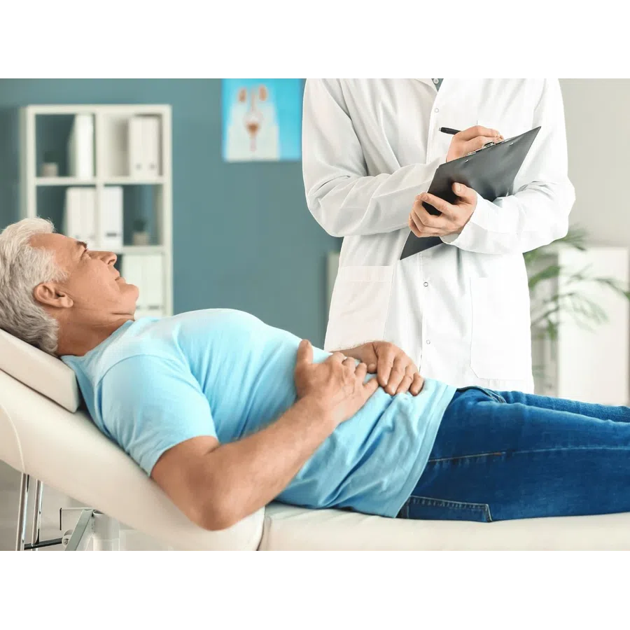Senior man lying on medical examination table holding abdomen while doctor takes notes in clinic room.