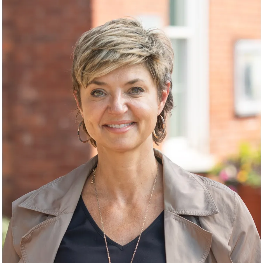 Smiling middle-aged woman with short blonde hair wearing a beige jacket and black top outdoors in soft light