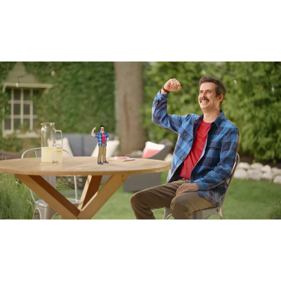 A man in a plaid shirt interacts with a miniature animated version of himself at a table.