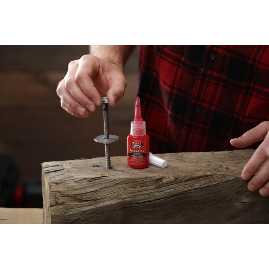 Hand applying red threadlocker adhesive to a metal bolt standing upright on a wooden surface with a plaid shirt background.