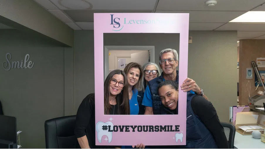 Five smiling dental staff posing behind a pink frame with #LOVEYOURSMILE at Levenson Smile clinic.