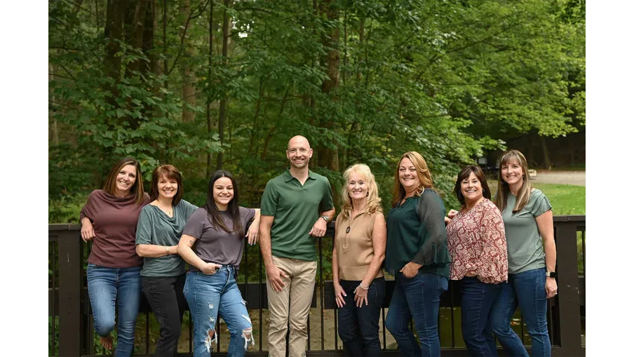 Group of eight adults posing outdoors on a wooden bridge with trees and greenery in the background