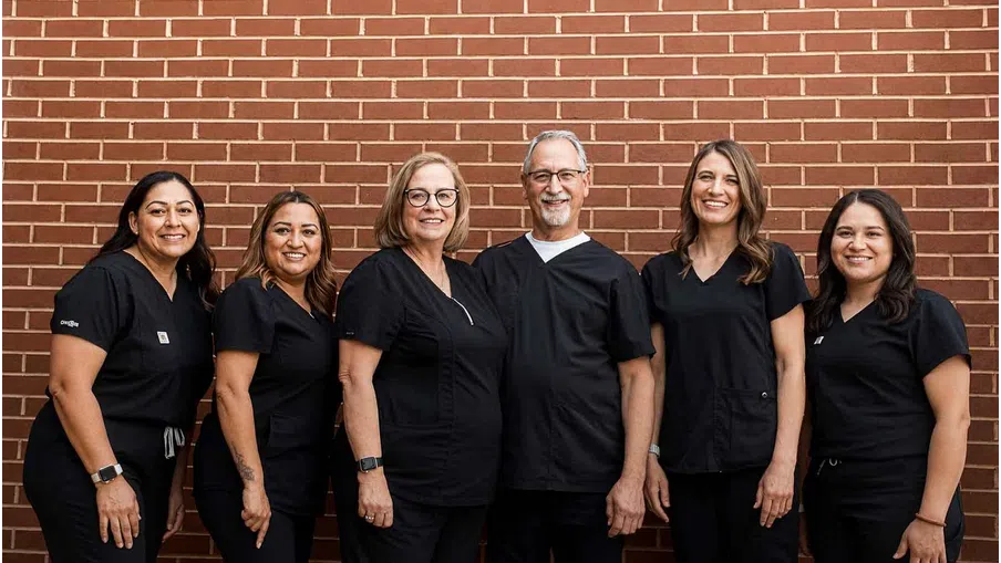 Group photo of six smiling healthcare professionals wearing black scrubs standing against a red brick wall.