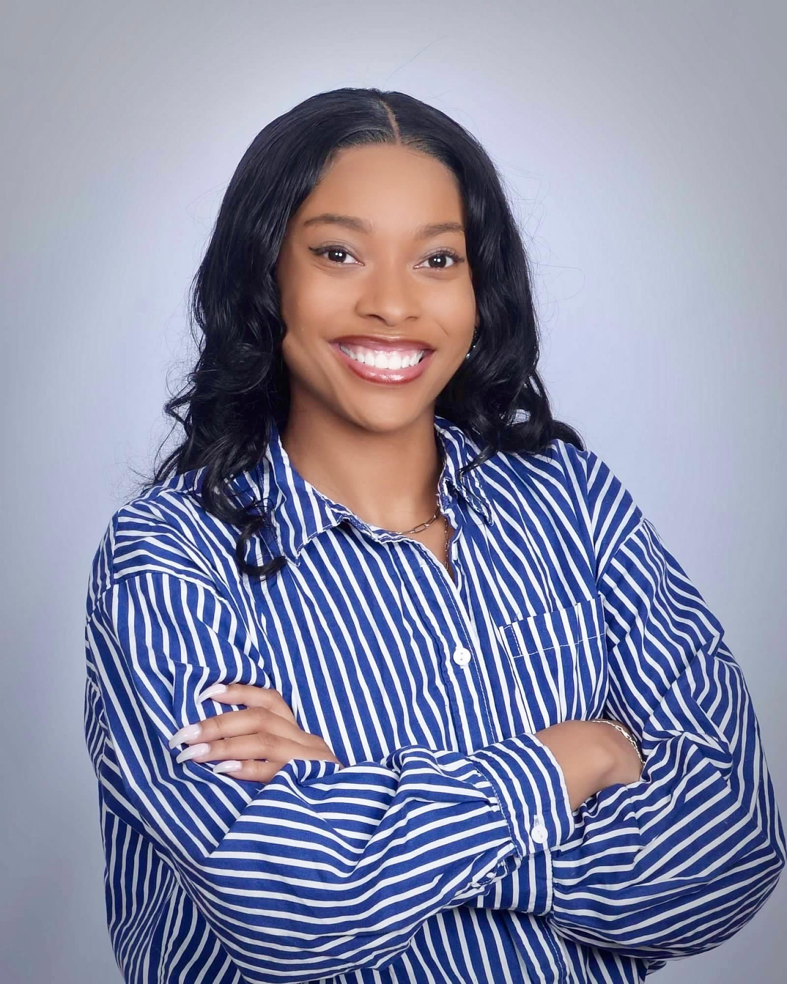 Smiling woman with long curly hair wearing a blue and white striped shirt standing with arms crossed.