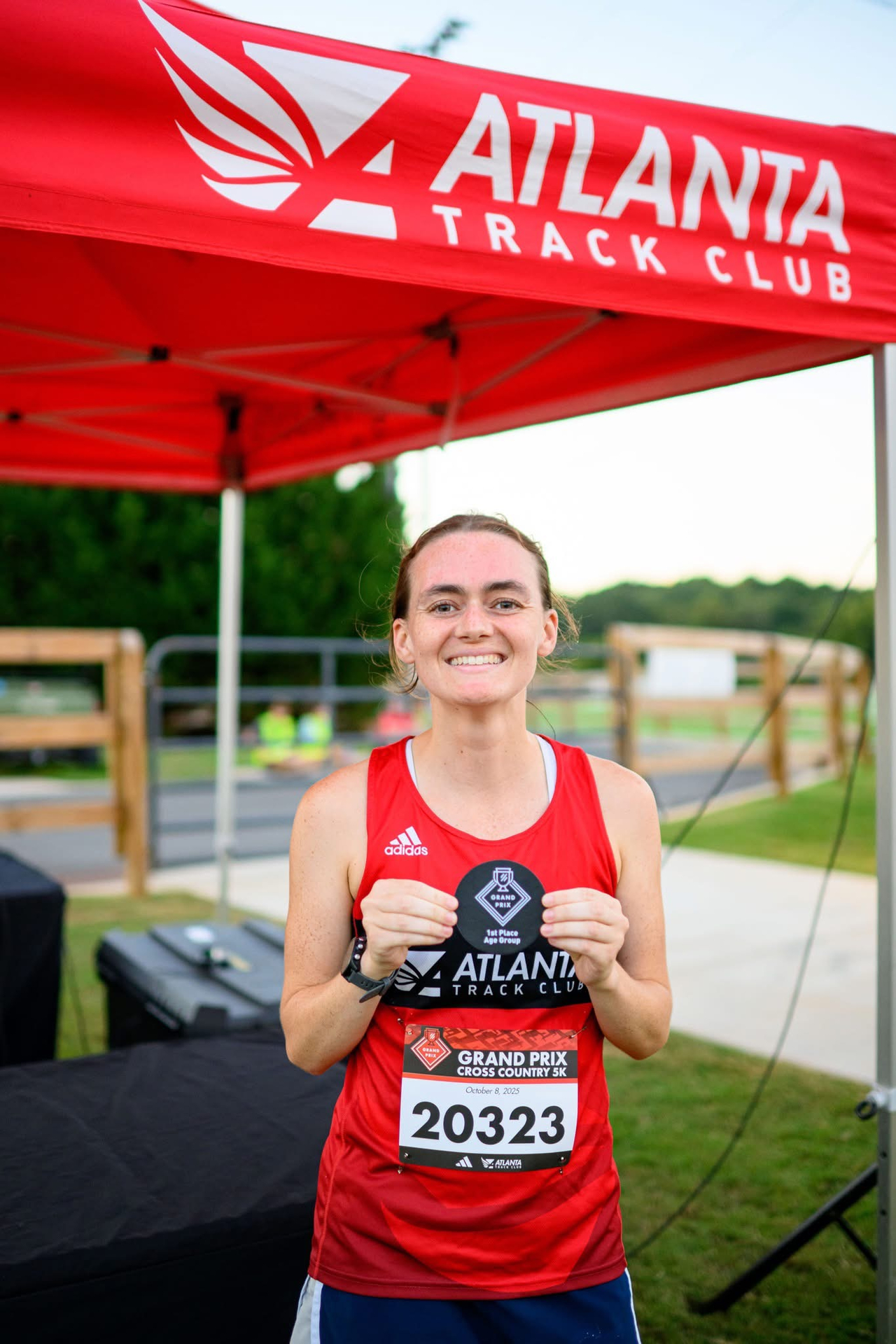 Smiling female runner in red Atlanta Track Club shirt holding first place medal at Grand Prix Cross Country 5K.